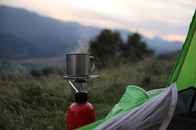 Boiling drink in metal mug on camping stove near tent outdoors
