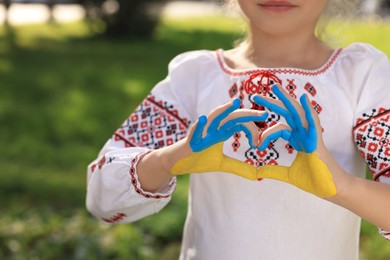 Little girl making heart with her hands painted in Ukrainian flag colors outdoors, closeup and space for text. Love Ukraine concept
