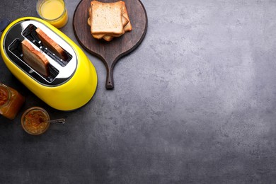 Yellow toaster with roasted bread, glass of juice and jam on grey table, flat lay. Space for text