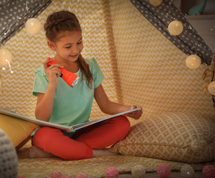 Photo of Little girl with flashlight reading book in play tent