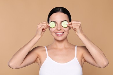 Beautiful young woman putting slices of cucumber on eyes against beige background