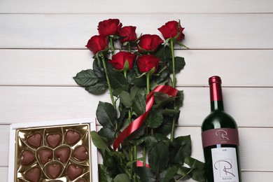 Photo of Bottle of red wine, beautiful roses and heart shaped chocolate candies on white wooden table, flat lay
