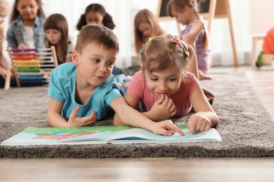 Cute kids reading book on floor while other children playing together in kindergarten