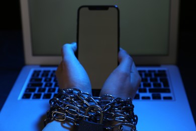 Woman with chained hands holding smartphone near laptop on dark background, closeup. Internet addiction