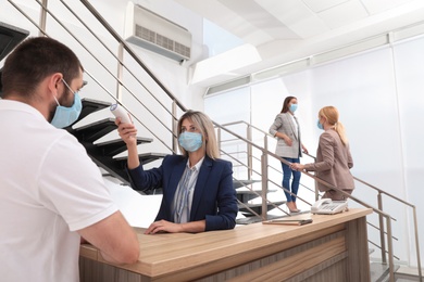 Photo of Woman in mask measuring temperature of employee with noncontact thermometer at office reception