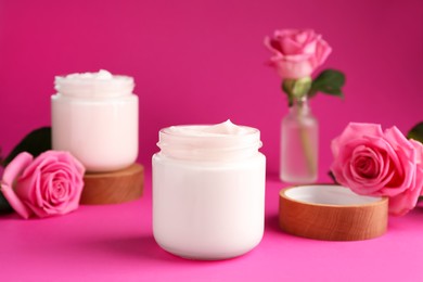 Glass jars of face cream and roses on pink background