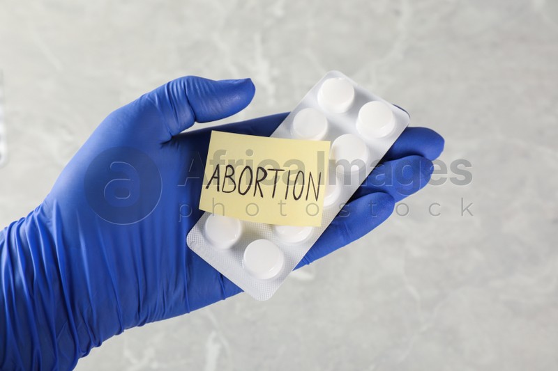 Doctor holding pills and paper card with word ABORTION on light grey background, closeup