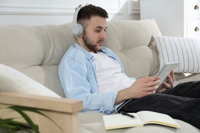 Young man with headphones using modern tablet for studying on sofa at home. Distance learning