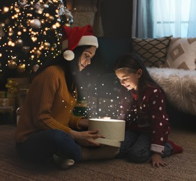 Mother with her cute daughter opening gift box with magical light at home. Christmas celebration