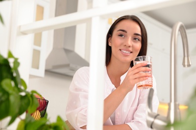 Young woman holding glass of pure water in kitchen, view through window