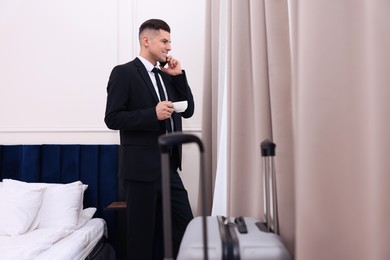 Handsome businessman drinking coffee while talking on phone near window in hotel room