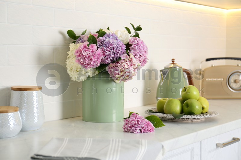 Beautiful hydrangea flowers, kettle and apples on light countertop