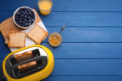 Yellow toaster with roasted bread, glass of juice, blueberries and jam on blue wooden table, flat lay. Space for text