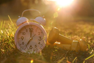 Photo of Alarm clock and dumbbells on green grass outdoors, closeup with space or text. Morning exercise