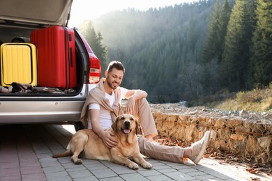 Photo of Happy man and adorable dog sitting near car in mountains. Traveling with pet
