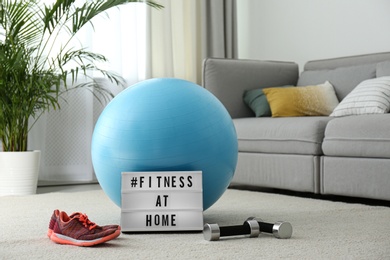 Sport equipment and lightbox with hashtag FITNESS AT HOME on floor indoors. Message to promote self-isolation during COVID‑19 pandemic