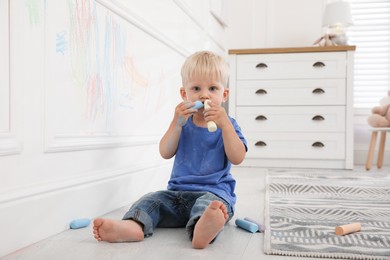 Mischievous little boy near white wall with colorful chalk scribbles at home