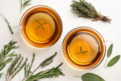 Cups of aromatic herbal tea with thyme, rosemary and sage on white marble table, flat lay
