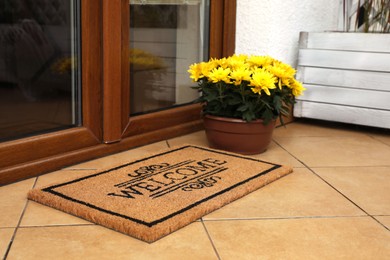 Door mat with word Welcome and beautiful flowers on floor near entrance