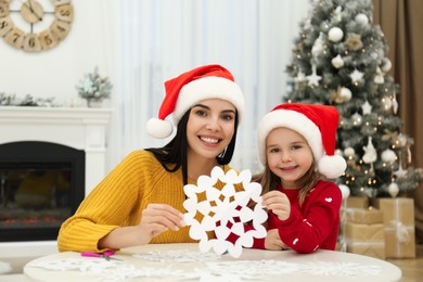 Mother and daughter in Santa hats with paper snowflake near Christmas tree at home