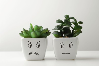 Beautiful potted houseplants with angry and concerned faces on white table. Emotional management