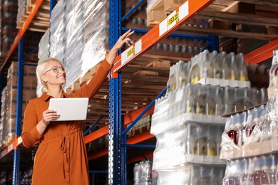 Photo of Happy manager holding modern tablet and pointing at something in warehouse with lots of products