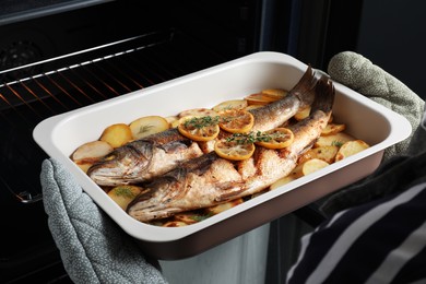Photo of Woman taking out baking tray with sea bass fish and potatoes from oven