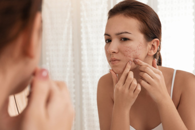 Photo of Teen girl with acne problem squeezing pimple near mirror in bathroom