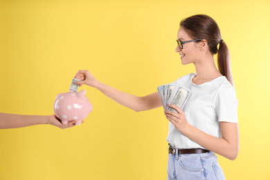Young woman putting money into piggy bank on yellow background