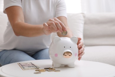 Photo of Woman putting coin into piggy bank at table indoors, closeup
