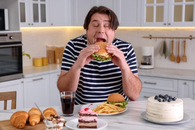 Hungry overweight man eating tasty burger at table in kitchen