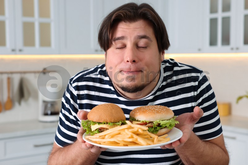 Happy overweight man holding plate with tasty burgers and French fries in kitchen