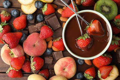 Fondue forks with strawberries in bowl of melted chocolate surrounded by other fruits on wooden table, flat lay