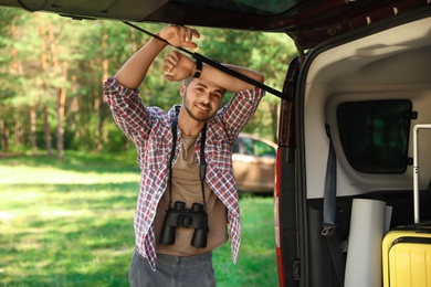 Young man with binoculars near van in forest. Camping gear