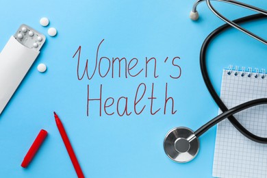 Photo of Words Women's Health, stethoscope, pills and stationery on light blue background, flat lay