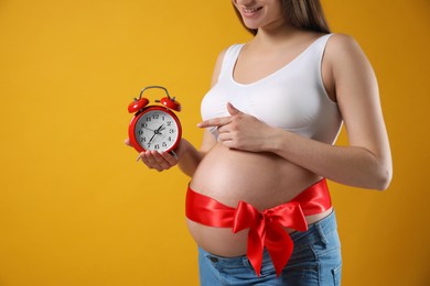Young pregnant woman with alarm clock and red bow on yellow background, closeup. Time to give birth