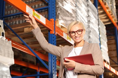 Photo of Happy manager holding modern tablet and pointing at something in warehouse with lots of products