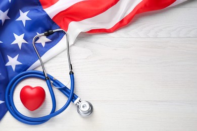 Photo of American flag, heart and stethoscope on white wooden table, flat lay. Space for text