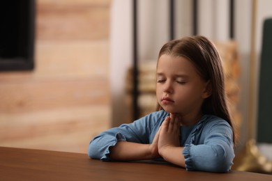 Cute little girl with hands clasped together praying at table indoors