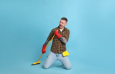 Handsome young man with floor brush singing on light blue background