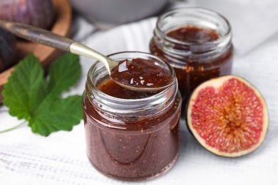 Photo of Glass jars of tasty sweet fig jam with spoon and fruits on white wooden table, closeup