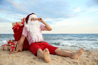 Photo of Santa Claus with bag of presents relaxing on beach, space for text. Christmas vacation