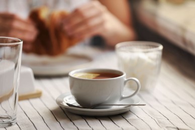 Woman eating croissant at table, focus on cup of aromatic tea with lemon