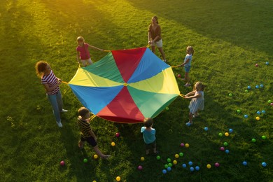 Group of children and teachers playing with rainbow playground parachute on green grass, above view. Summer camp activity