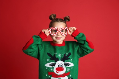 Cute little girl in Christmas sweater and party glasses on red background