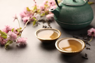 Photo of Traditional ceremony. Cups of brewed tea, teapot and sakura tree branch on grey table, closeup