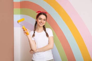 Young woman holding roller near wall with painted rainbow indoors