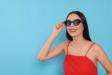 Attractive happy woman touching fashionable sunglasses against light blue background. Space for text