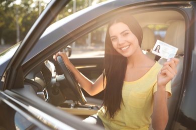 Happy young woman holding license while sitting in car outdoors. Driving school