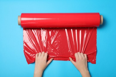 Photo of Woman unrolling red plastic stretch wrap on light blue background, top view